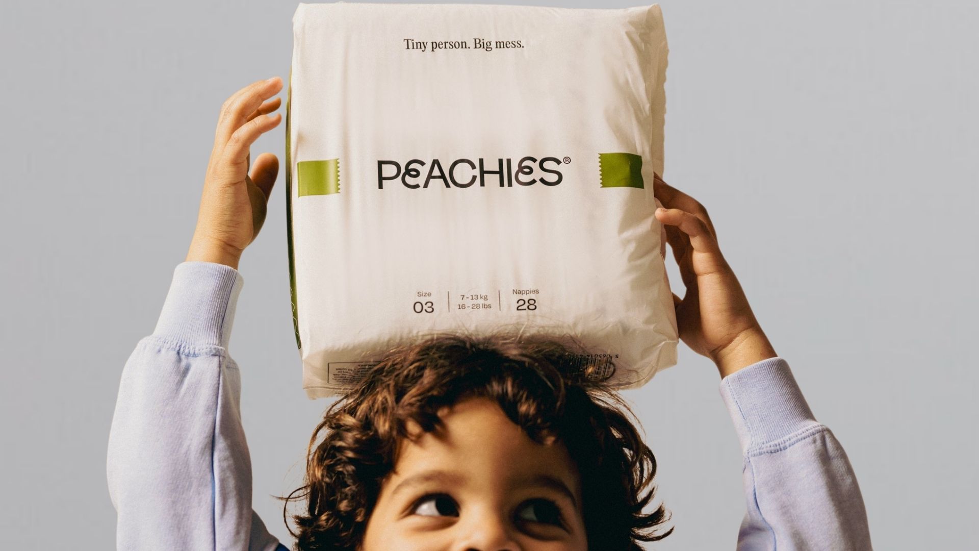 A close-up shot of a young child with dark, curly hair looking slightly upward while balancing a large white pack of Peachies nappies on their head. The child’s hands are raised on either side to steady the package. The background is a clean, neutral light grey.