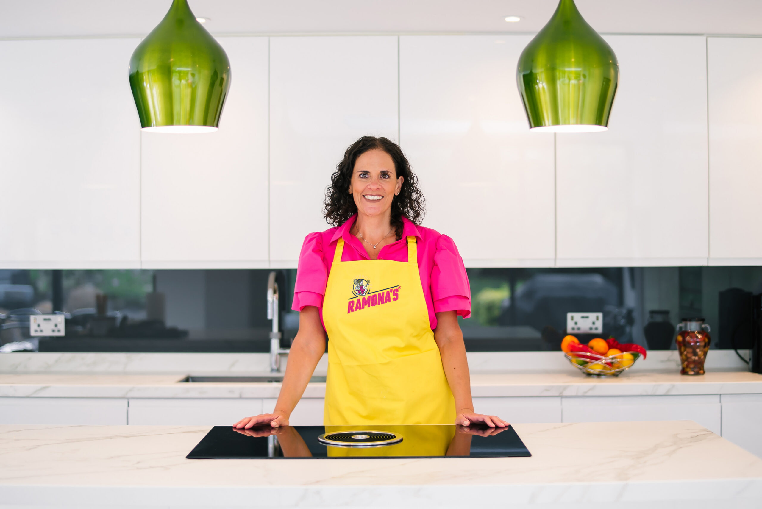 RAMONA-New Brand – Smile A female entrepreneur wearing a yellow Ramona's apron stands in a modern white kitchen, representing food business support at Imperial Enterprise Lab.